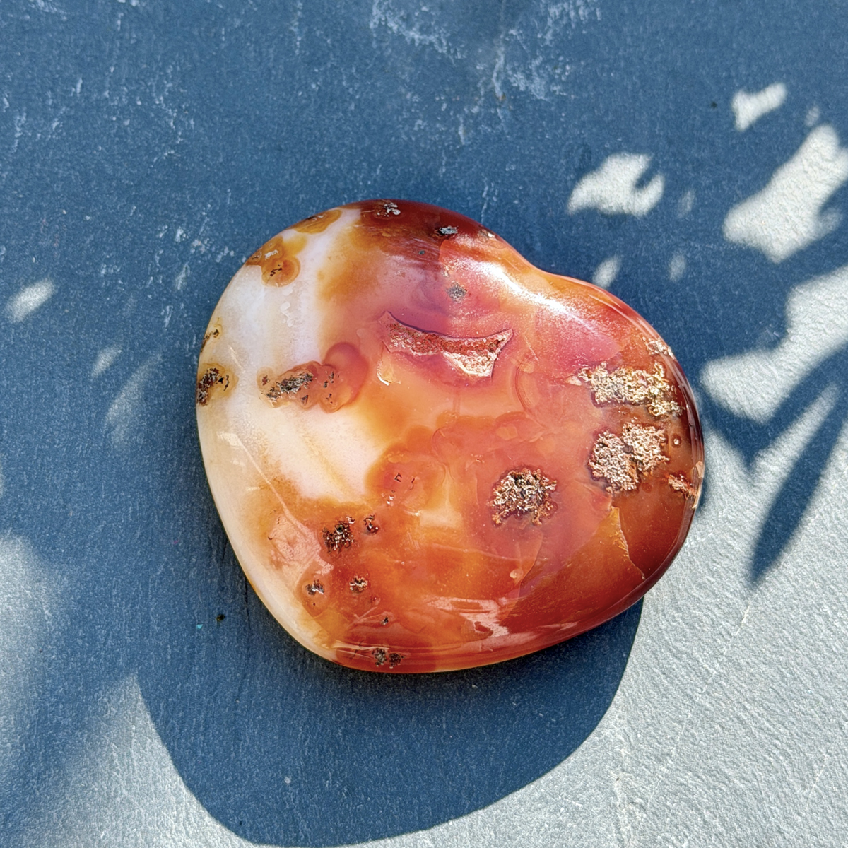 Coeur en cornaline naturelle de Madagascar posé sur fond bleu texturé, face principale visible avec ses nuances rouge-orangé intenses et ses inclusions minérales dorées. Pierre dynamisante lithothérapie, idéal comme cadeau bien-être original.