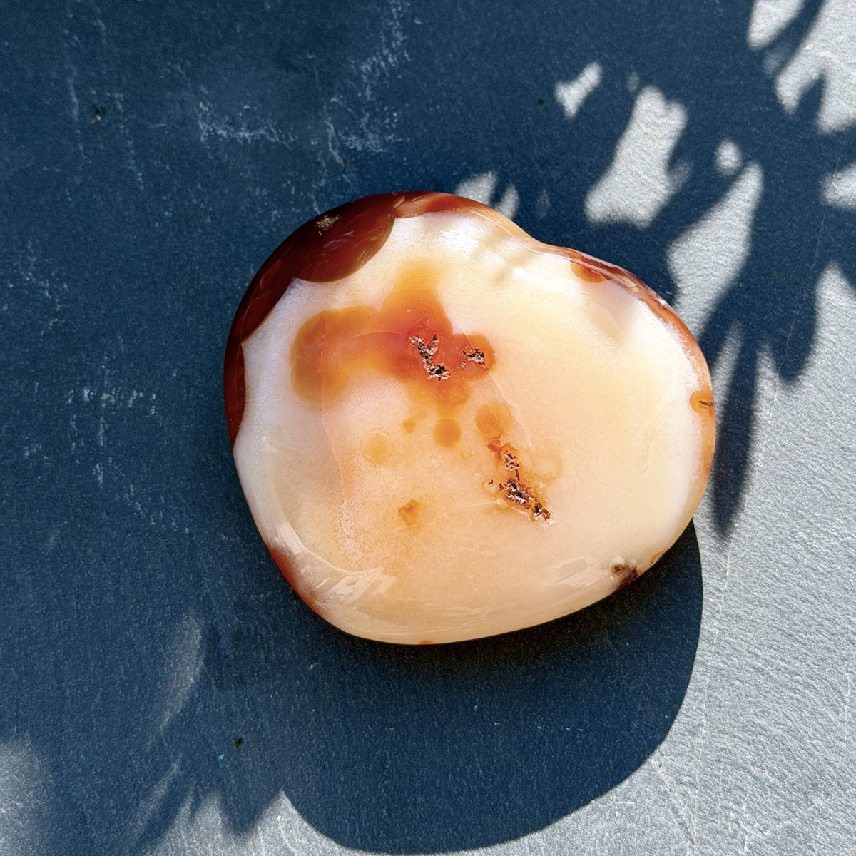 Vue de profil d'un coeur en cornaline naturelle aux tons blanc crème et orangé, posé sur fond bleu en pleine lumière. Les inclusions minérales caractéristiques de la cornaline de Madagascar sont visibles en détail. Idée cadeau lithothérapie pour femme.