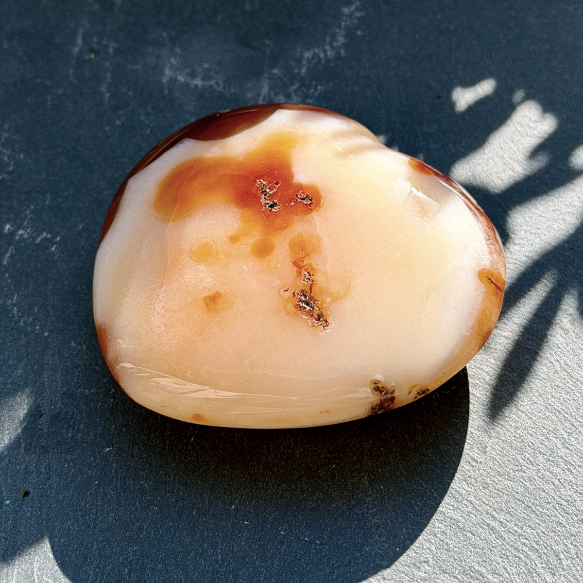 Coeur en cornaline naturelle photographié de face en lumière directe sur fond bicolore bleu et gris. Les teintes rouge, orange et rosé de la pierre brillent intensément. Pierre de la vitalité et de l'élan créatif en lithothérapie. Cadeau bien-être femme.