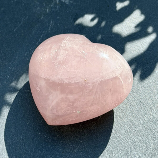 coeur-quartz-rose-polie-vue-profil-soleil Vue de profil d'un coeur en quartz rose poli aux reflets nacrés, photographié en lumière naturelle sur fond bleu texturé. La forme bombée et généreuse de la pierre est mise en valeur. Objet de lithothérapie, cadeau original pour femme.