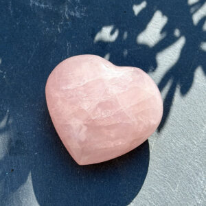 Coeur en quartz rose naturel photographié en pleine lumière sur un fond bicolore bleu et gris clair, avec jeu d'ombres douces. Pierre de douceur et d'amour inconditionnel en lithothérapie. Idée cadeau bien-être et spiritualité.