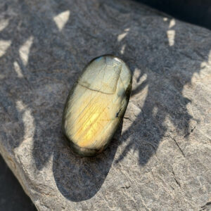 Vue du dessus d'un galet en labradorite naturelle ambrée poli, posé à l'angle d'une roche granitique en plein soleil. Les reflets or et ambre chauds de la labradorescence sont intenses et lumineux. Pierre rare lithothérapie, cadeau bien-être original. Belle de Lune Bijoux.