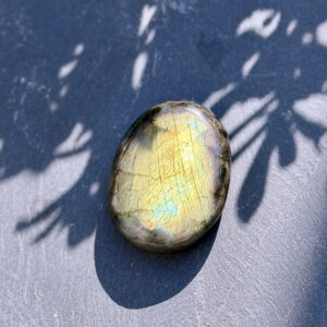 Galet en labradorite naturelle aux reflets jaune doré et turquoise posé sur fond bleu bicolore avec ombres de feuillage projetées. La labradorescence multicolore est particulièrement lumineuse en plein soleil. Pierre naturelle non traitée, idéale pour la méditation. Belle de Lune Bijoux.