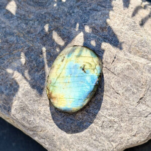 Vue du dessus d'un galet en labradorite naturelle poli, posé sur une pierre minérale en plein soleil. Les reflets bleu et or caractéristiques de la labradorescence sont parfaitement visibles. Pierre de protection et d'intuition en lithothérapie. Belle de Lune Bijoux.