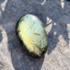 galet-labradorite-dessus-reflets-vert-or-roche-soleil Vue du dessus d'un galet en labradorite naturelle poli aux dominantes vert et or, posé sur une roche granitique en plein soleil avec jeu d'ombres. Pierre de protection et de transformation en lithothérapie. Belle de Lune Bijoux.