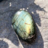 galet-labradorite-face-vert-dore-roche-ombre-feuillage Galet en labradorite naturelle de Madagascar photographié de face sur une roche minérale, entouré d'ombres de feuillage. Les reflets vert intense et dorés de la labradorescence sont parfaitement visibles. Pierre lithothérapie pour la clarté mentale et l'intuition. Belle de Lune Bijoux.