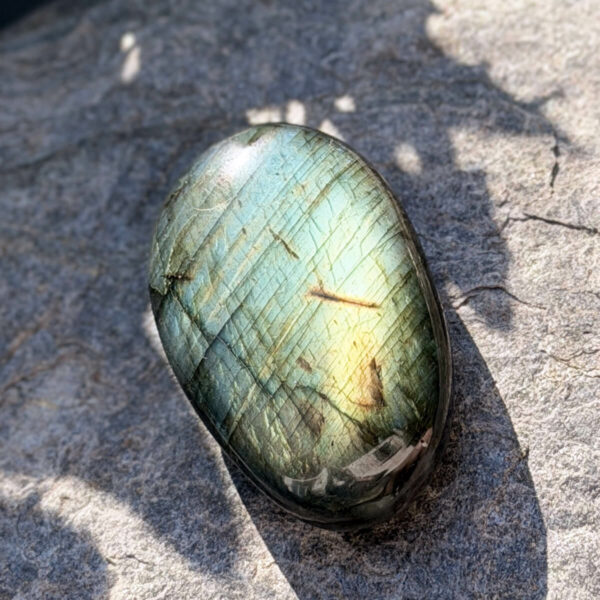 galet-labradorite-face-vert-dore-roche-ombre-feuillage Galet en labradorite naturelle de Madagascar photographié de face sur une roche minérale, entouré d'ombres de feuillage. Les reflets vert intense et dorés de la labradorescence sont parfaitement visibles. Pierre lithothérapie pour la clarté mentale et l'intuition. Belle de Lune Bijoux.