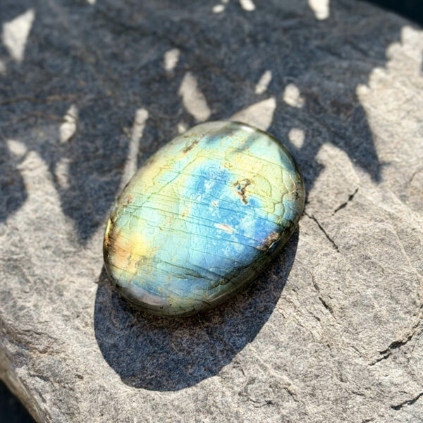 galet-labradorite-main-reflets-bleu-jaune-fond-vert Galet en labradorite naturelle de Madagascar tenu dans une main ouverte, photographié en extérieur sur fond de végétation floue. Les reflets bleu intense et jaune doré de la labradorescence illuminent la surface polie. Pierre de protection énergétique idéale pour les personnes sensibles.