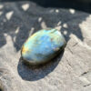 galet-labradorite-pierre-reflet-orange-dore-lumiere-soleil Galet en labradorite naturelle posé sur une pierre brute en lumière directe, révélant un reflet orange et doré spectaculaire en bas de la pierre. La labradorescence aux tons chauds et irisés est particulièrement visible. Pierre rare lithothérapie, cadeau bien-être original.