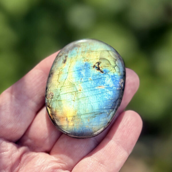 galet-labradorite-polie-vue-face-roche-ombre-feuillage Vue de profil d'un galet en labradorite naturelle polie, posé à l'angle d'une roche granitique en plein soleil. Les reflets bleu-vert et dorés de la surface sont mis en valeur par la lumière naturelle. Pierre de l'intuition et de la protection, idéale pour la méditation.