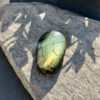 galet-labradorite-vue-face-roche-reflets-vert-jaune-soleil Galet en labradorite naturelle posé sur une roche en lumière directe, face principale visible avec ses nuances vert clair et jaune doré caractéristiques. La labradorescence aux tons chauds est particulièrement lumineuse. Pierre naturelle non traitée, idéale en méditation.